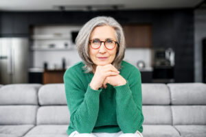 gray-haired female with hands on chin in glasses sitting on couch in living room at home, inquisitively looking at camera