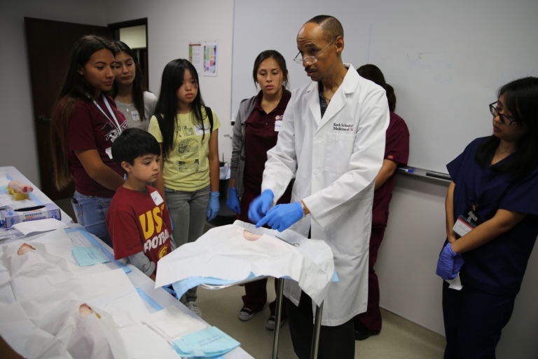 Man in white coat demonstrates suturing techniques to group of students learning about health care careers.