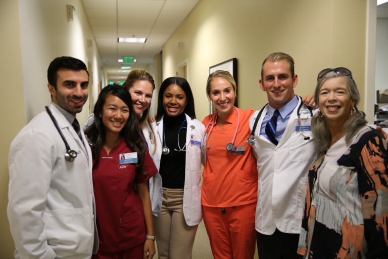 Group of PA students in white coats stand with faculty in clinical setting.