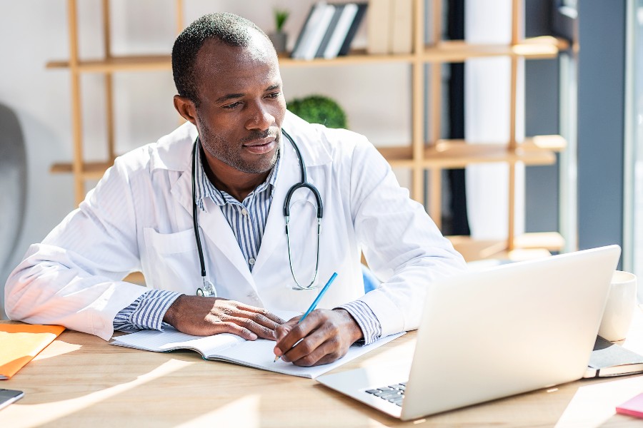 Physician looks at his laptop while taking notes.