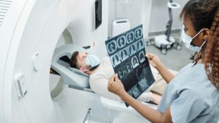 Young African radiologist in uniform and protective mask looking at x-ray image of patient lying on long couchette of medical equipment