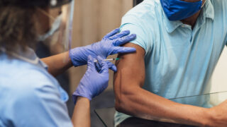 A mixed race female nurse wearing a protective face shield, surgical mask and protective gloves administering the COVID-19 vaccine to a senior black man in his home.