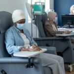 Two senior women sit side-by-side in their own chairs as they receive their cancer treatments intravenously. They are both dressed casually and have head scarves on as they sit looking across the room. A nurse can be seen working at a desk in the background.
