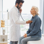 Side view of focused adult man in white coat using stethoscope while elderly woman sitting on exam couch. General practitioner examining heartbeat while providing regular checkup in clinic.