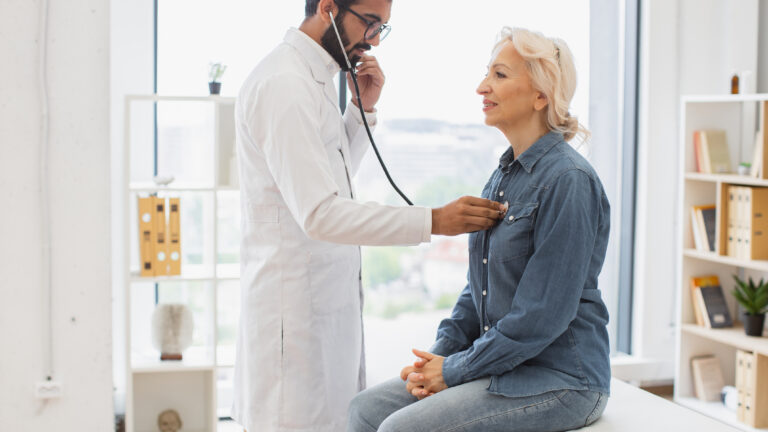 Side view of focused adult man in white coat using stethoscope while elderly woman sitting on exam couch. General practitioner examining heartbeat while providing regular checkup in clinic.