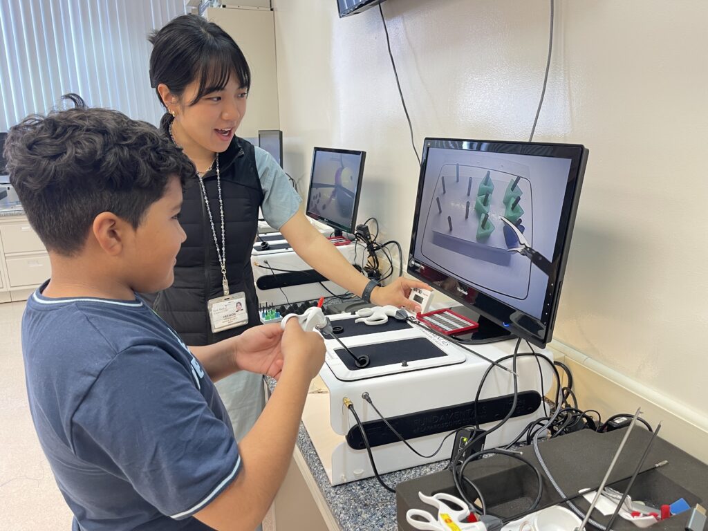 Medical student shows middle-schooler how to use a machine simulating laparoscopic and robotic procedures.