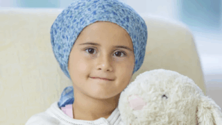 A little girl wearing a blue headscarf holds a stuffed animal, smiling at the camera.