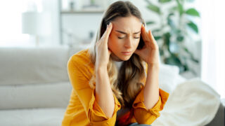 Stressed young woman in a yellow shirt sitting on a couch at living room, pressing her fingers to her temples with a pained expression, having migraine