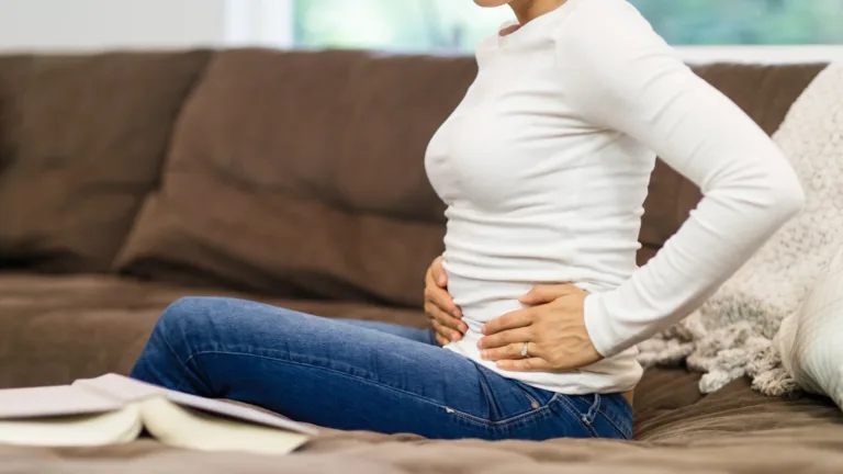 A woman sitting on a couch, holding her stomach, showing signs of discomfort and pain.