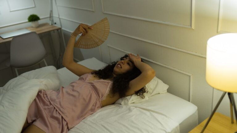 Photo shows young woman lying in bed awake fanning herself, unable to sleep.