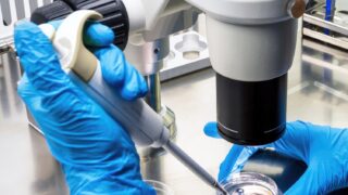 A high-resolution microscope with a gloved hand holding a pipette.