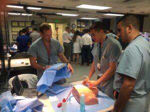 Three medical students in green scrubs practice surgical techniques on a partial dummy