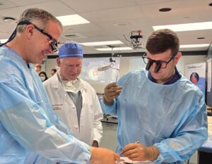 Two surgeons in protective gear work on a practice procedure. Dr. Vaughn Starnes, in the middle, is observing, He wears a white surgeon's jacket and a surgical cap.