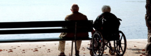 Elderly couple seen from behind at the beach. The woman is in a wheelchair.